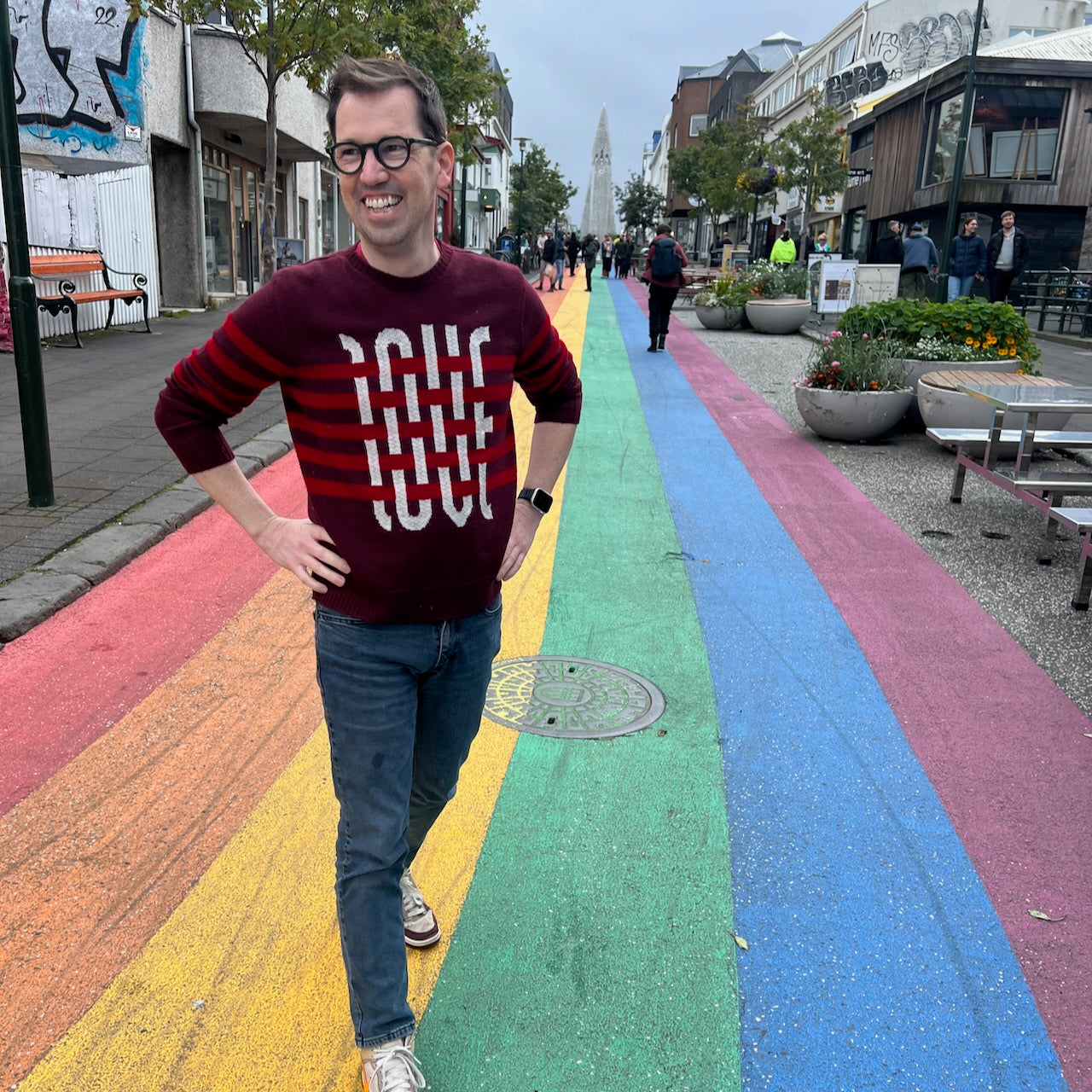 Jedd standing on a rainbow painted road wearing a sweater that says Love, and the Reykjavik Cathedral in the background